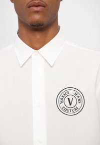 Chemise à col blanc en tissu lisse, avec un logo circulaire noir portant "VERSACE JEANS COUTURE" et un design central en forme de "V".