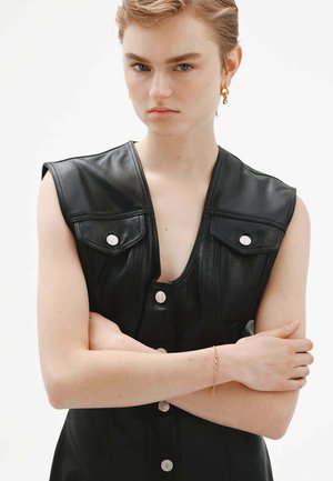 Vestido sin mangas de cuero negro con un profundo escote en V, que presenta botones de presión plateados y bolsillos en el pecho, colocado sobre un fondo neutro.