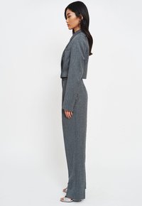 Szary dopasowany garnitur z krótką marynarką i szerokimi spodniami. Teksturowana tkanina i subtelny wzór. Srebrne sandały na obcasie. Widok z boku.