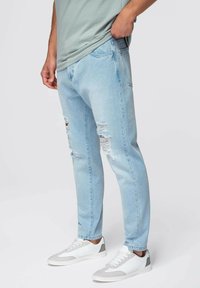 Jeans de mezclilla color azul claro con detalles desgastados. Diseño ajustado con pierna cónica, combinados con zapatillas blancas y grises.