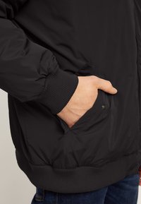 Black insulated jacket with a smooth texture, ribbed cuffs, and a side pocket with a snap closure. Casual design with elastic hem.