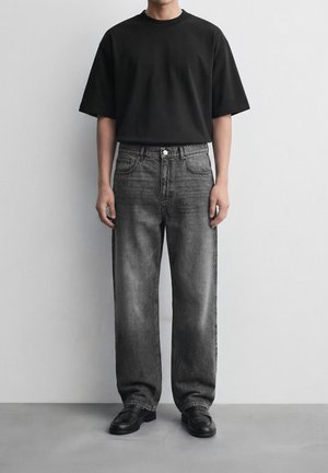 Άτομο που φοράει φαρδιά μαύρη κοντομάνικη μπλούζα, ξεθωριασμένο γκρι τζιν με φαρδιά πόδια και μαύρα παπούτσια, στέκεται μπροστά σε έναν απλό λευκό τοίχο.