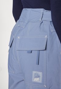 Light blue cargo pants with multiple pockets, visible white stitching, and a branded patch. Features adjustable waistband and smooth texture.