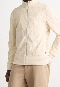 Man i en krämfärgad, texturerad zip-up-jacka med ribbade manschetter och nederkant, i kombination med beigefärgade byxor, stående med en hand i fickan.