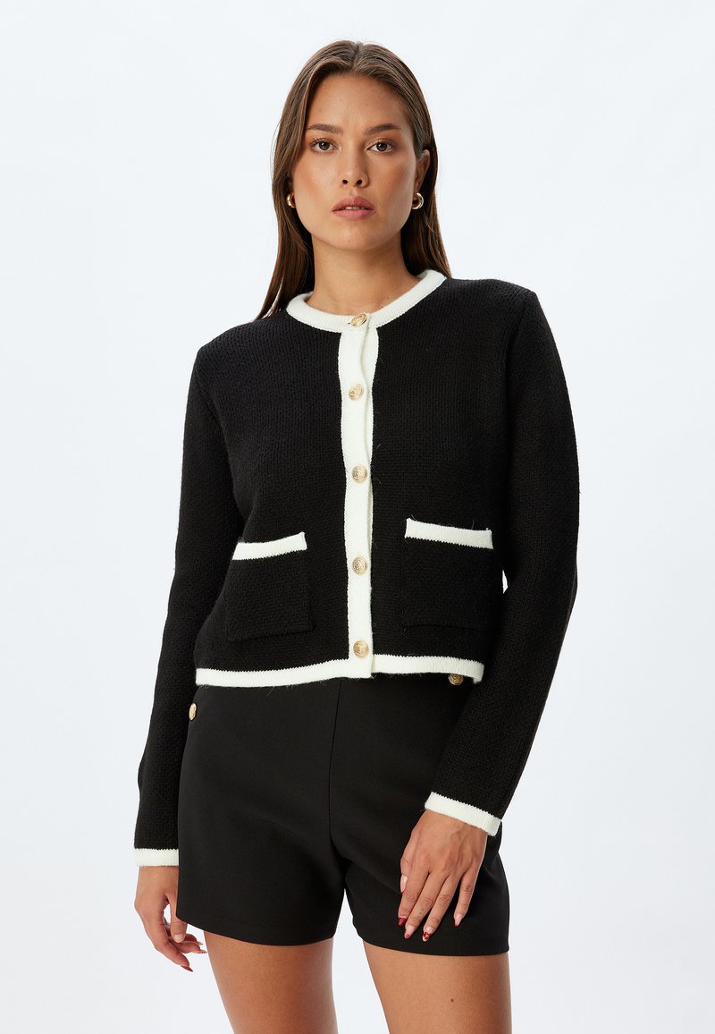 Svart cardigan med krämfärgad kant, rund halsringning och fyra guldknappar. Har två framfickor. Texturerad tyg. Korth design.