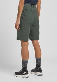 Dunkelgrüne Shorts mit glatter Textur, knielangem Schnitt, hohem Bund und flacher Vorderseite. Getragen mit schwarzen Schuhen und grauen gemusterten Socken.
