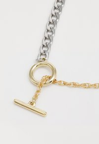 Cadena de plata gruesa que lleva a un anillo de metal dorado con una cadena dorada y cierre de barra, con acabados pulidos y colores contrastantes.