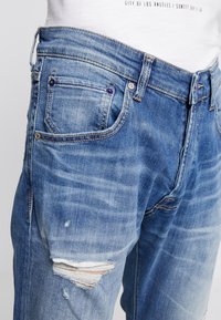 Jeans de denim azul con un lavado descolorido, detalles desgastados y un diseño clásico de cinco bolsillos. Presenta costuras doradas y acentos de hardware metálico.