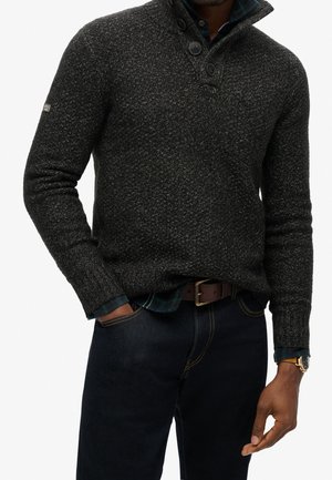 Homme portant un pull à col boutonné gris foncé texturé par-dessus une chemise à carreaux verte et un jean foncé, les mains dans les poches.