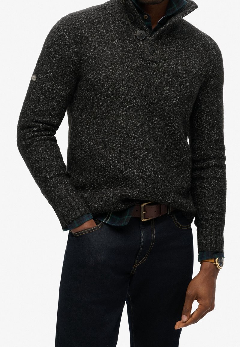 Homme portant un pull à col boutonné gris foncé texturé par-dessus une chemise à carreaux verte et un jean foncé, les mains dans les poches.