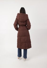 Abrigo de plumas marrón con capucha, cintura ceñida y diseño acolchado. Combinado con jeans azules de pierna acampanada y zapatos de tacón transparente.