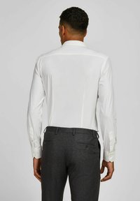 Chemise blanche à manches longues avec un col standard, en tissu lisse, et poignets boutonnés, associée à un pantalon texturé gris foncé avec des poches arrière.