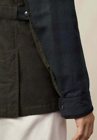 Tamnozelena i mornarskoplava jakna s rukavom na kojem je dugme, koja miruje u blizini ruke, uparena s krem hlačama.