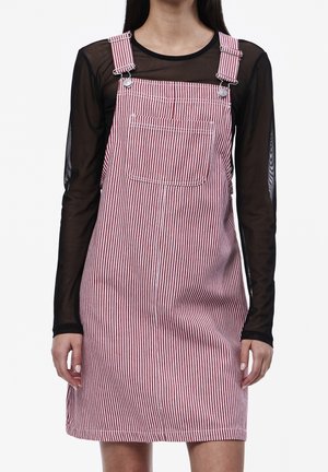 Personne portant une robe salopette rayée rouge et blanche avec des bretelles réglables sur un haut noir transparent à manches longues, sur un fond uni.