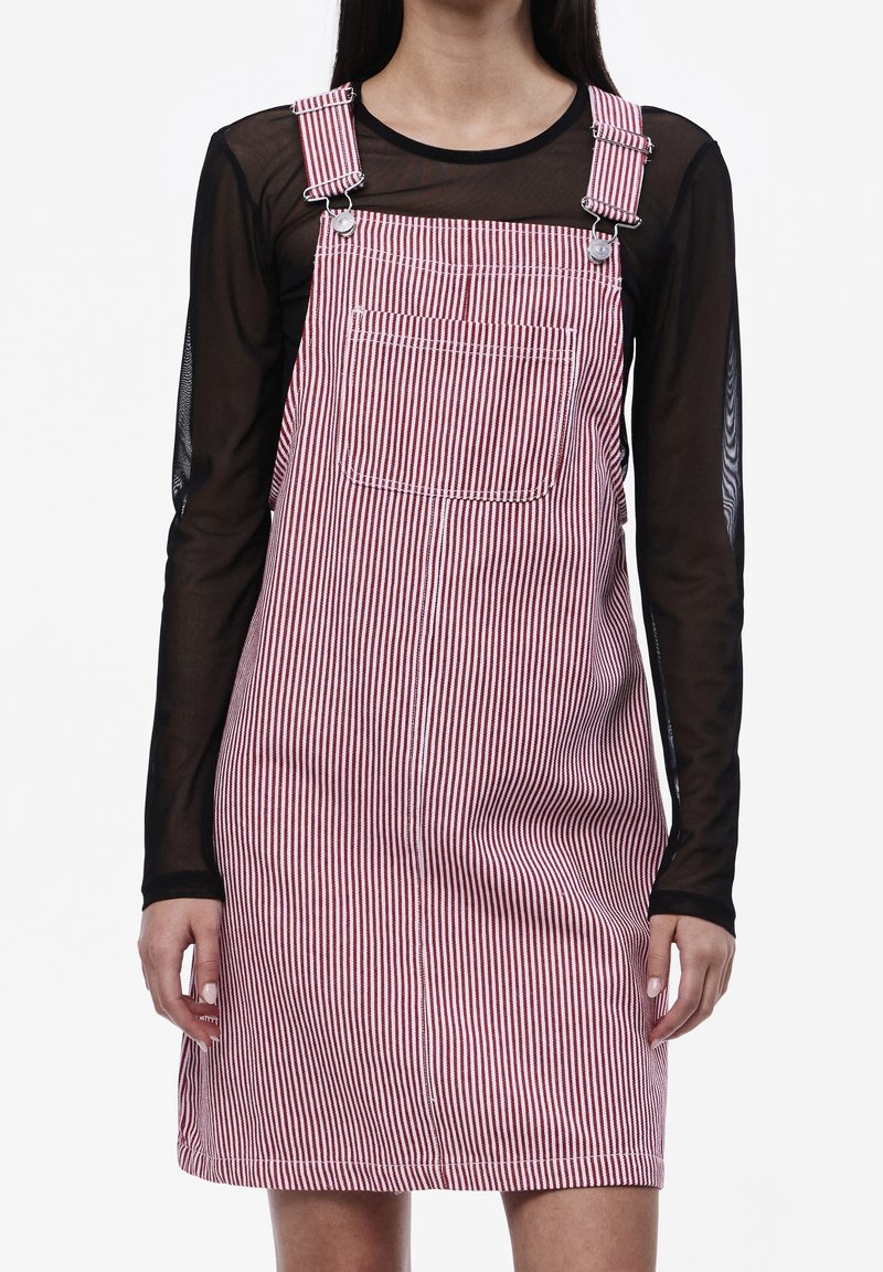 Personne portant une robe salopette rayée rouge et blanche avec des bretelles réglables sur un haut noir transparent à manches longues, sur un fond uni.