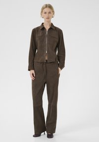 Bruine leren jas met ritssluiting, twee borstzakken en bijpassende high-waisted pantalons. Glad textuur, op maat gesneden pasvorm en uitlopende pijpen.