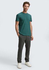 Turkusowa bawełniana koszulka z okrągłym dekoltem, zestawiona z ciemnoszarymi teksturowanymi spodniami i białymi sneakersami, stojąca na jasnym tle.