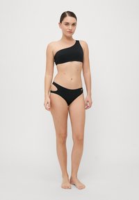 Μαύρο μπικίνι με ένα ώμο και κοψίματα στο κάτω μέρος, φτιαγμένο από απαλό ύφασμα με ελάχιστα αξεσουάρ.