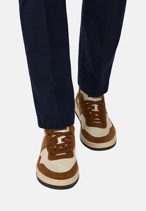 Braune und cremefarbene Sneaker aus Wildleder und Leder; mit runden Schnürsenkeln, perforiertem Zehenbereich und kontrastierender Sohle. Getragen mit navyfarbigen Hosen.