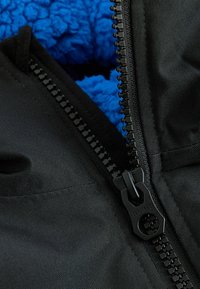 Svart yttertyg med en synlig svart dragkedja som visar en texturerad blå fleecefoder under. Närbild på material och dragkedjedetalj.