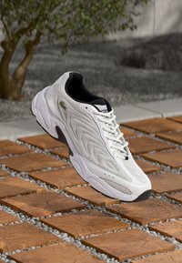 Fehér sneaker texturált felsőrésszel, fekete részletekkel és kiemelkedő logóval. Felszerelt fűzős zárással és párnázott talppal rendelkezik. Könnyű kialakítás, a föld felett lebeg.