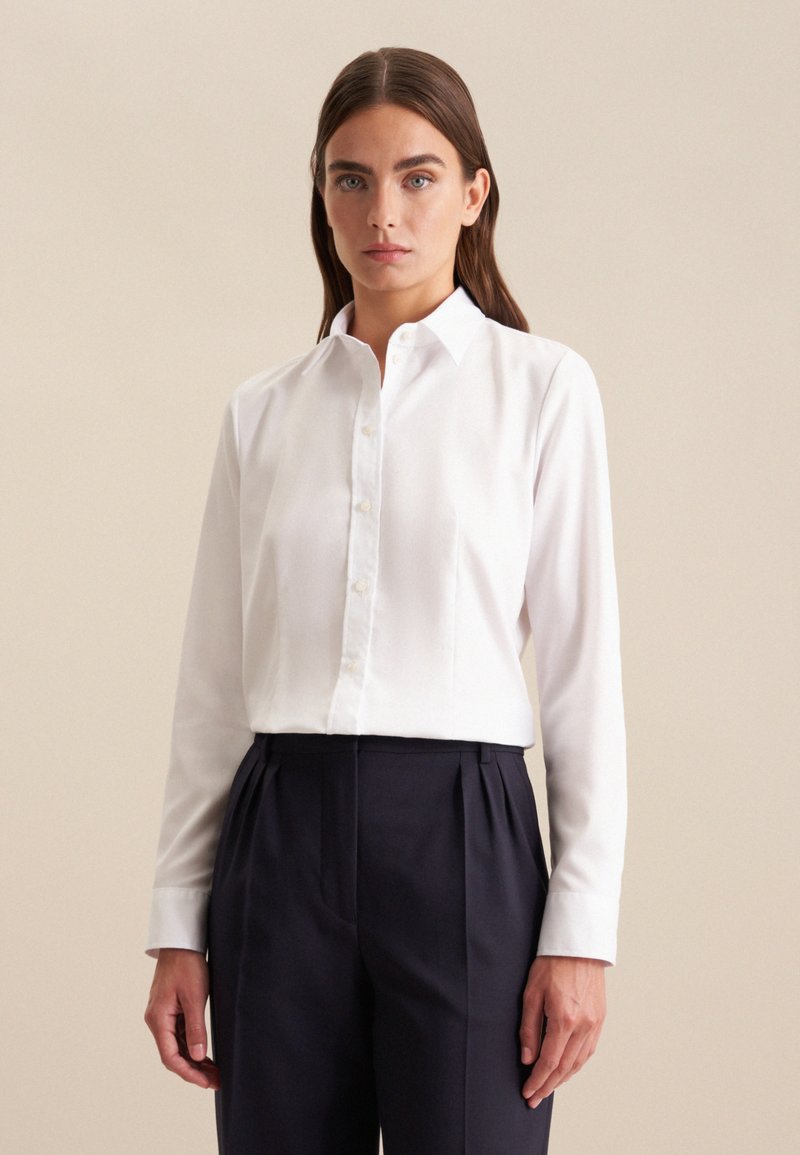 Camicia bianca con bottoni, realizzata in tessuto satinato, con colletto, maniche lunghe e vestibilità aderente, abbinata a pantaloni scuri.