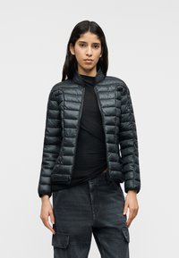 Μαύρο μπουφάν με οριζόντια ραμμένα πάνελ, ψηλό γιακά και πλήρες εμπρόσθιο φερμουάρ, φορεμένο πάνω από μαύρο μπλουζάκι και σκούρο παντελόνι.