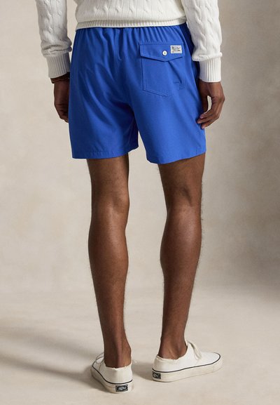 Homme portant un short bleu vif avec une poche arrière et des chaussures blanches à enfiler, debout devant un fond neutre.