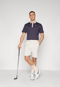 Sötétkék pólóing világos rózsaszín gallérral, bézs rövidnadrággal, fehér sportcipővel szürke részletekkel, és egy golfütővel, semleges háttér előtt.