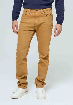 Jean homme couleur sable à coupe droite, cinq poches et tissu résistant. Stylé avec un pull bleu marine et des baskets blanches.