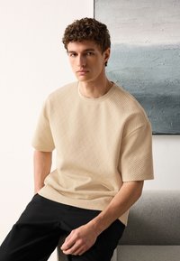 Beige textured short-sleeve sweater with a round neckline, paired with dark pants. Smooth fabric with a subtle geometric pattern.