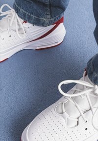 Scarpe da ginnastica bianche con dettagli rossi. Tessuti in rete e lisci, tomaia perforata, suole spesse. Indossate con jeans in denim blu su una superficie morbida.