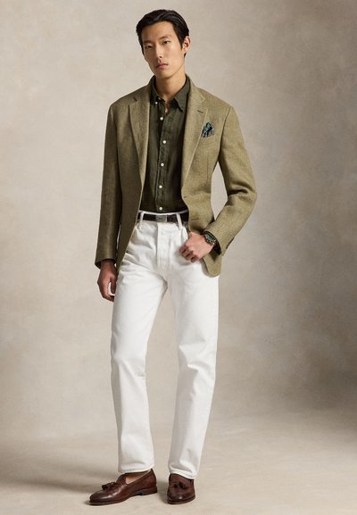 Férfi olíva színű blézerben, sötétzöld ingben, fehér nadrágban, barna rojtos mokaszinekben és zsebkendővel, áll egy egyszínű bézs háttér előtt.