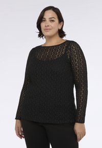 Žena s krátkými tmavými vlasy, oblečená v černém krajkovém topu s dlouhými rukávy a černých kalhotách, stojící proti jednoduchému světlému pozadí.