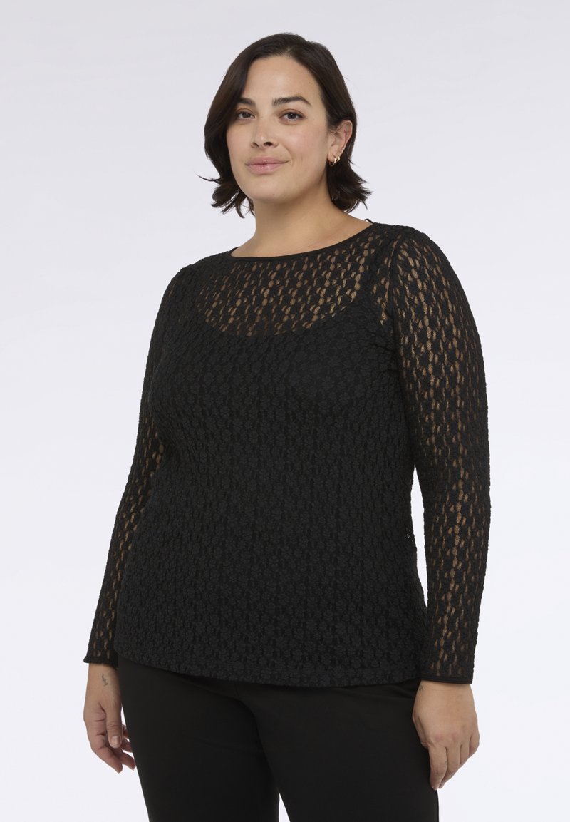 Žena s krátkými tmavými vlasy, oblečená v černém krajkovém topu s dlouhými rukávy a černých kalhotách, stojící proti jednoduchému světlému pozadí.