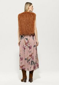 Braune, fransige, kurz geschnittene Pelz-Weste über einem langen, blumigen rosa Kleid mit lila und grünen Farbtönen; braune knielange Stiefel mit Absatz.