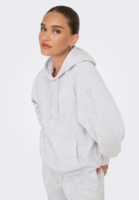 Felpa con cappuccio grigia chiara, dal taglio morbido e rilassato, con cappuccio regolabile e tasca frontale. Tessuto morbido e texture al tatto; abbinata a pantaloni della tuta grigi coordinati.