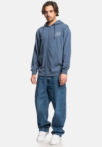 Modrá mikina na zips s predným vreckom a logom na hrudi, doplnená voľnými modrými džínami a bielymi teniskami.
