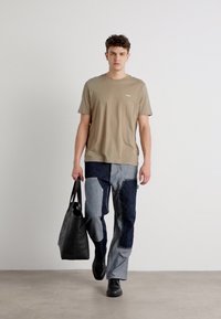 Beige t-shirt med rund hals, lappe-lomme, blå denim patchwork bukser, sorte sko og en sort tote taske. Ensfarvet baggrund, model der går.