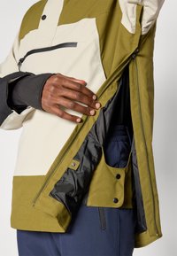 Oliiviv roheline ja beež jakk, millel on esi tõmblukuga tasku, reguleeritavad mansetid ja nööbiga sisetasku. Tekstuuriga kangas.