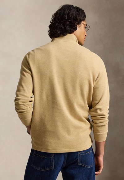 Homme aux cheveux bouclés et portant des lunettes, vêtu d'un pull beige à manches longues et d'un jean bleu, vu de dos sur un fond neutre.