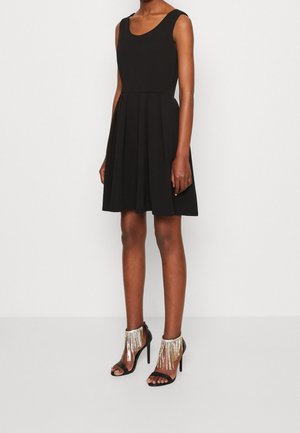 Robe noire sans manches avec un corsage ajusté et une jupe évasée. Associée à des talons noirs ornés de franges argentées autour de la cheville.