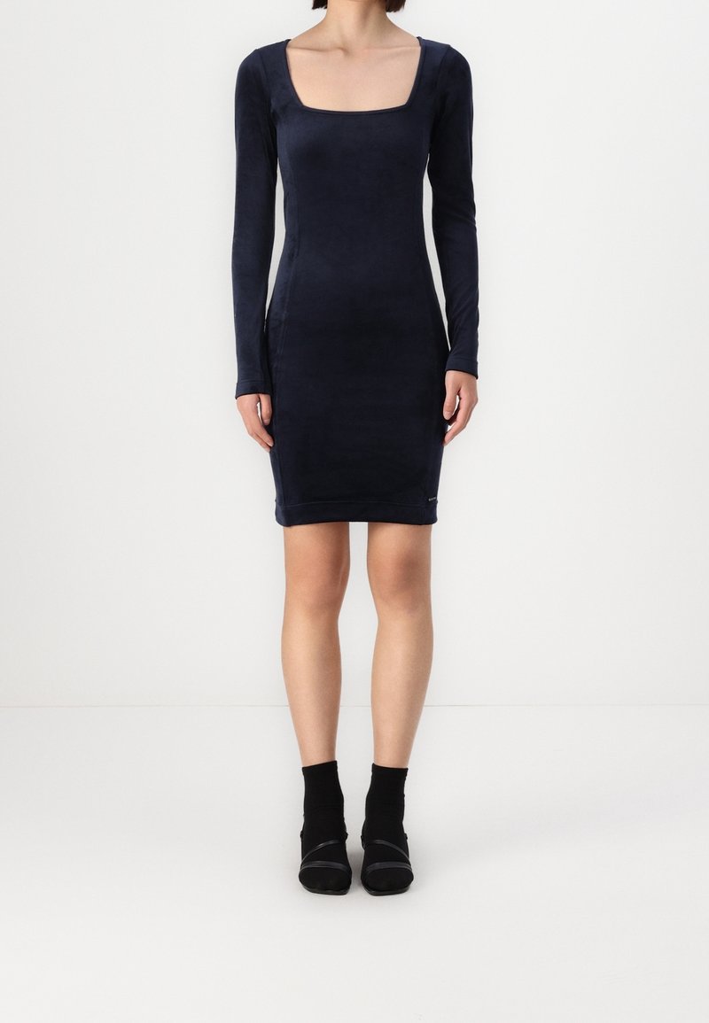 Femme portant une robe bleu marine ajustée à manches longues avec un col carré, associée à des chaussettes noires aux chevilles et des talons à bouts ouverts, debout devant un fond blanc.