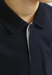 Mörkblå polo med en texturerad krage och vit detalj längs knäppningen, som visar upp ett mjukt tyg och strukturerade designelement.