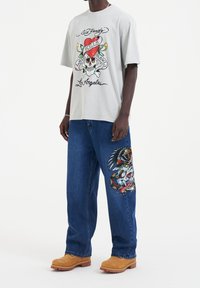 Szary t-shirt z kolorową grafiką czaszki i serca; niebieskie dżinsy z detalicznym haftowanym wzorem; brązowe buty; luźny krój.