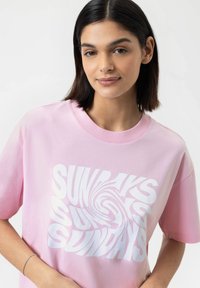 T-shirt en coton rose avec un col rond et des manches courtes. Présente un texte blanc en spirale audacieux lisant "DIMANCHES" dans un design ludique.