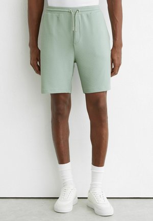 Mężczyzna ubrany w jasnooliwkowe szorty z teksturą i sznurkiem, białe skarpetki do kostki oraz białe trampki na jednolitym białym tle.