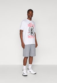 Witte grafische t-shirt met roze en zwarte print, gecombineerd met grijze shorts. Model draagt witte sportschoenen en sokken met zwarte accenten.