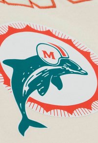 Ein Delfin, der einen weißen Helm mit einem roten "M" trägt, innerhalb eines weißen Kreises, umgeben von einem roten Sonnenstrahlenmuster auf einem beigen Hintergrund.