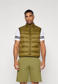 Olivgrüne Pufferweste mit hohem Kragen, Reißverschluss und schwarzen Akzenten, getragen über einem weiß gestreiften T-Shirt und olivgrünen Shorts.